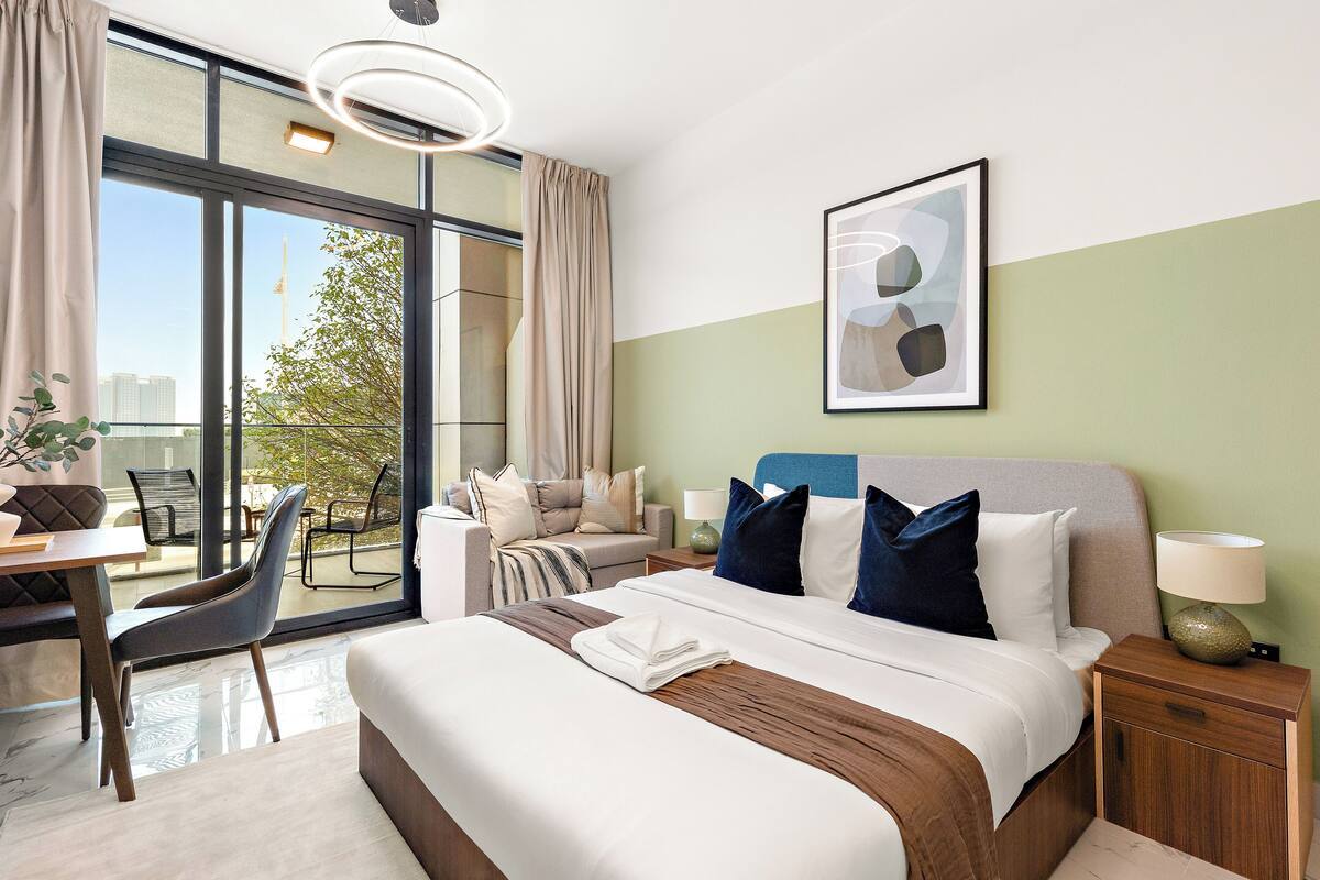 A hotel room with a large bed, a desk, a chair, a balcony with a view of buildings and trees, and a framed abstract painting on the wall.