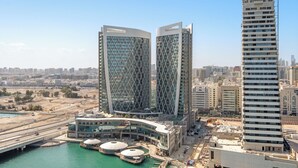 A modern urban area with two prominent buildings featuring a geometric design, a waterfront with a bridge, and a mix of commercial and residential structures. - Silkhaus Al Maryah Vista 1 (Abu Dhabi)