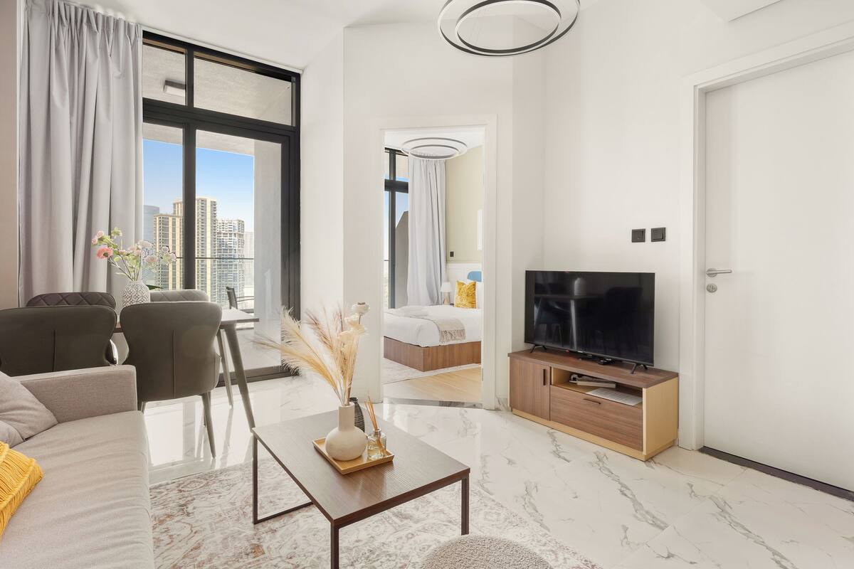 A modern hotel room with a large window, a flat-screen TV, a sofa, a coffee table, and a bed with a view of the cityscape.