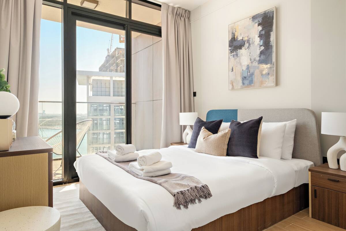 A modern hotel room with a large bed, a view of the city, and a balcony with a chair.