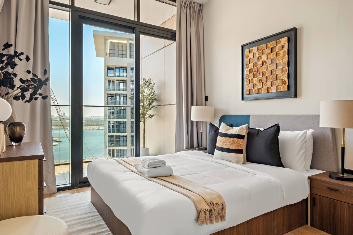 A modern hotel room with a large bed, a view of the city, and a balcony with a plant.