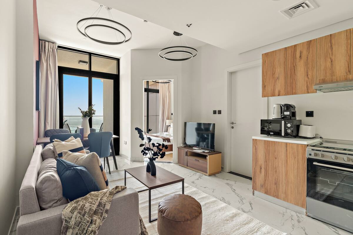A modern living room with a sofa, coffee table, and a TV. There's a kitchen with wooden cabinets and a stove.