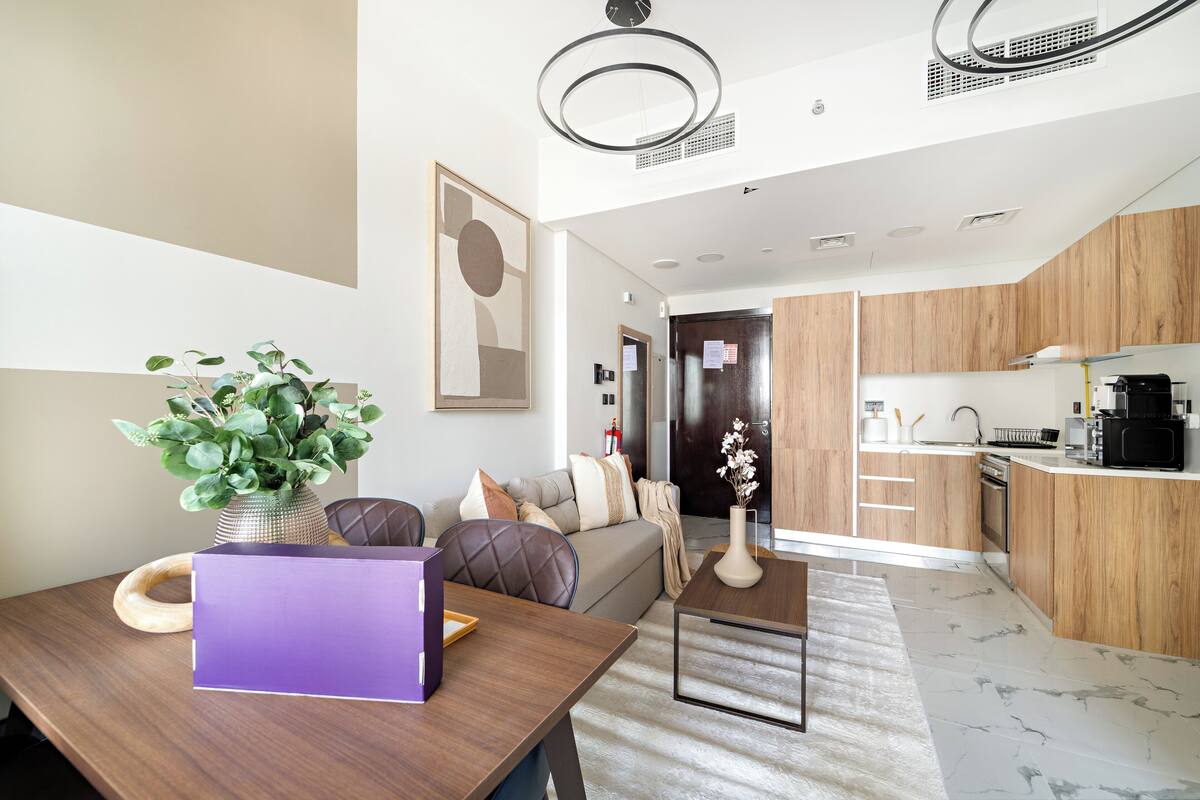 A modern living room with a sofa, a coffee table, and a dining table. There is a kitchen area with wooden cabinets and a microwave.