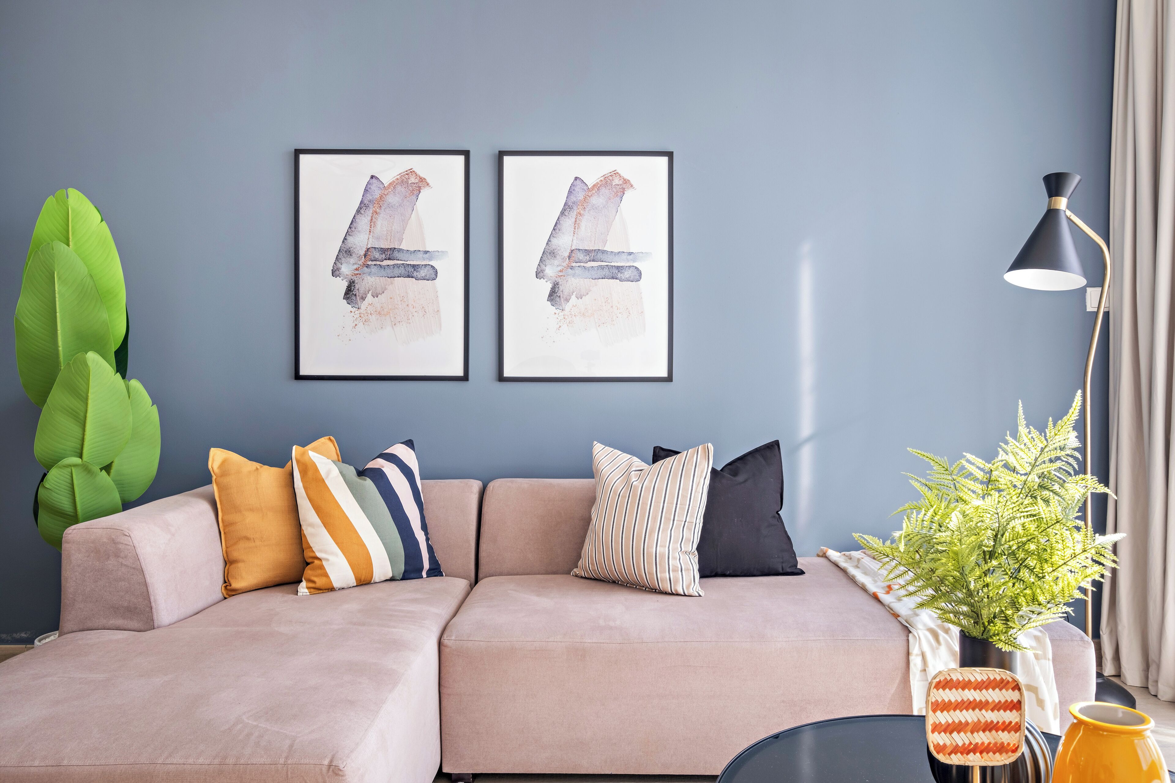 A modern living room with a light-colored sectional sofa, two framed abstract paintings, a green plant, and a floor lamp.