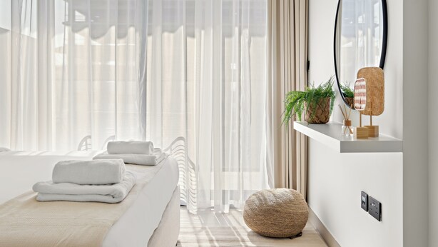A modern bedroom with a large window, a bed with neatly folded towels, a round woven ottoman, and a wall-mounted mirror.