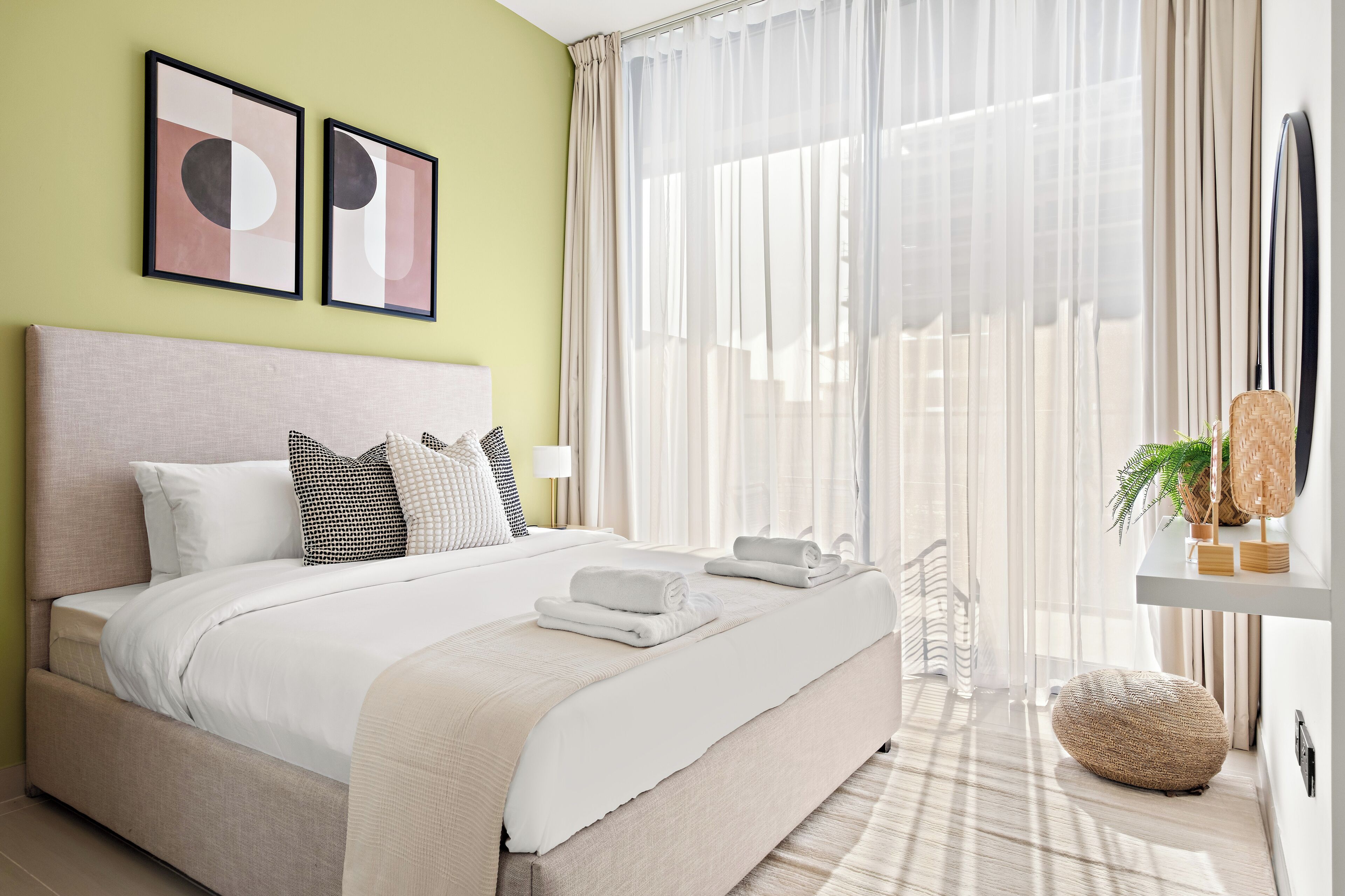 A modern bedroom with a large bed, a bedside table, a potted plant, and a view of the outdoors through sheer curtains.