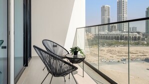 A balcony with a glass railing, two black chairs, a small table with a plant, and a view of tall buildings and a parking lot.