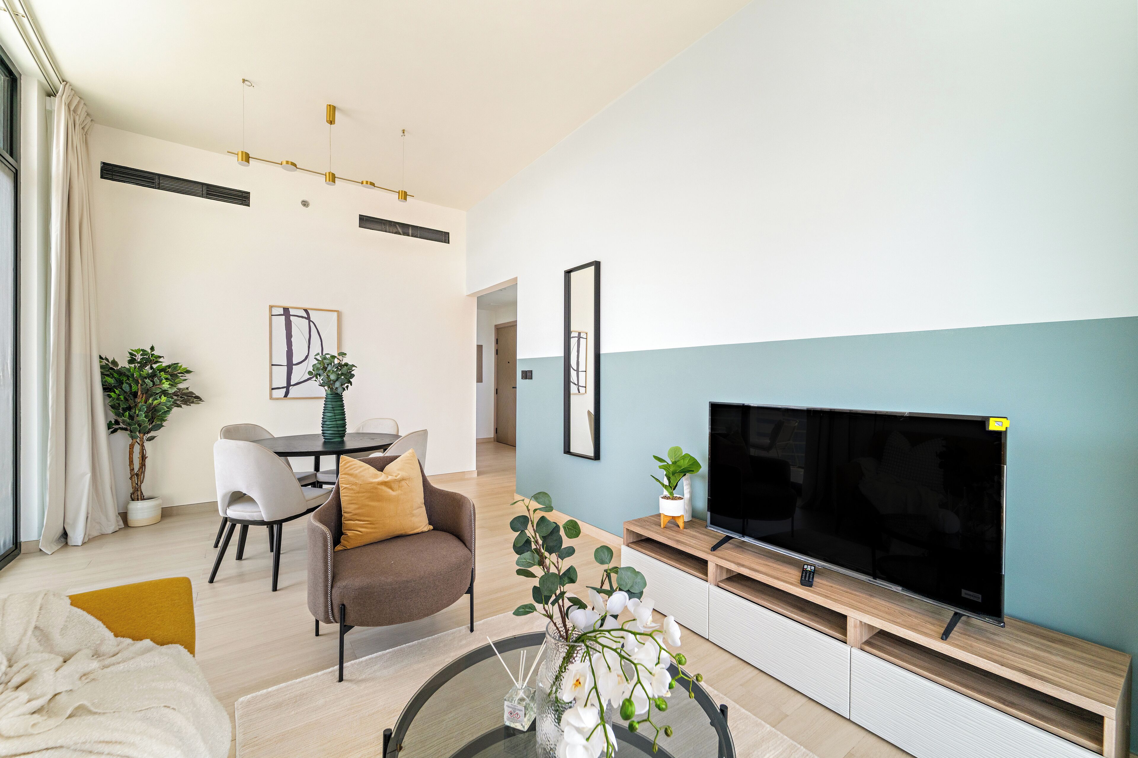 Uma sala de estar moderna com televisão de tela plana, área de jantar com cadeiras, sofá e mesa de centro com um vaso de flores.