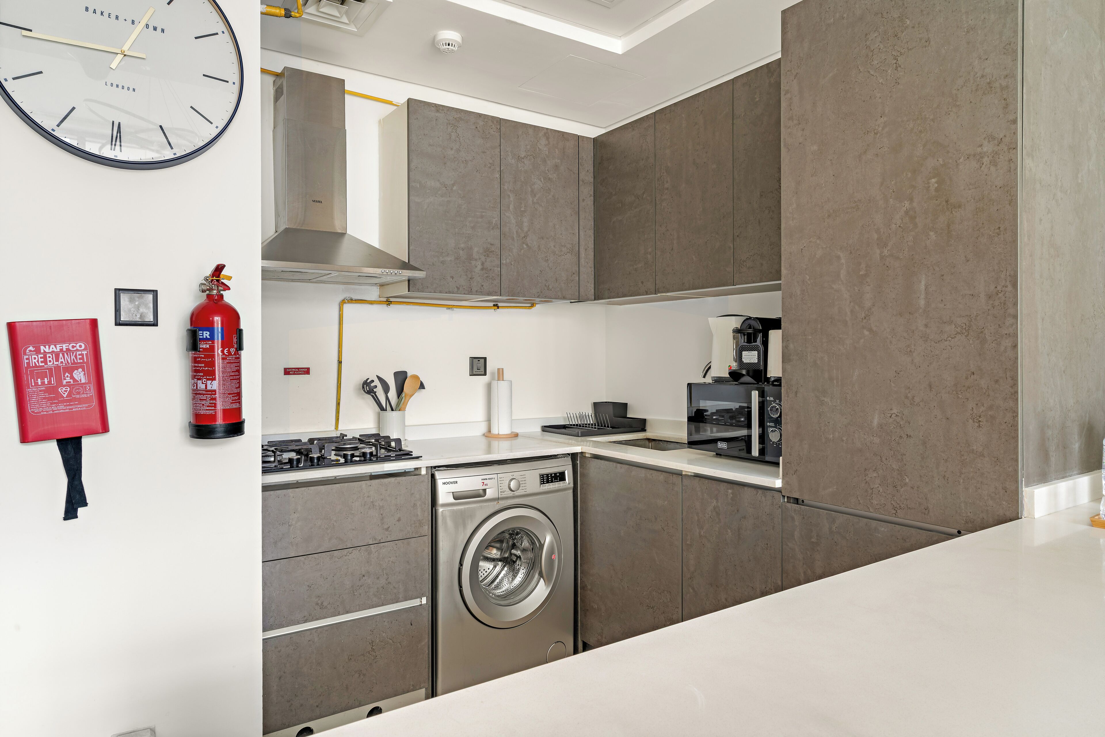 A modern kitchen with grey cabinets, a washing machine, a fire extinguisher, and a clock on the wall.