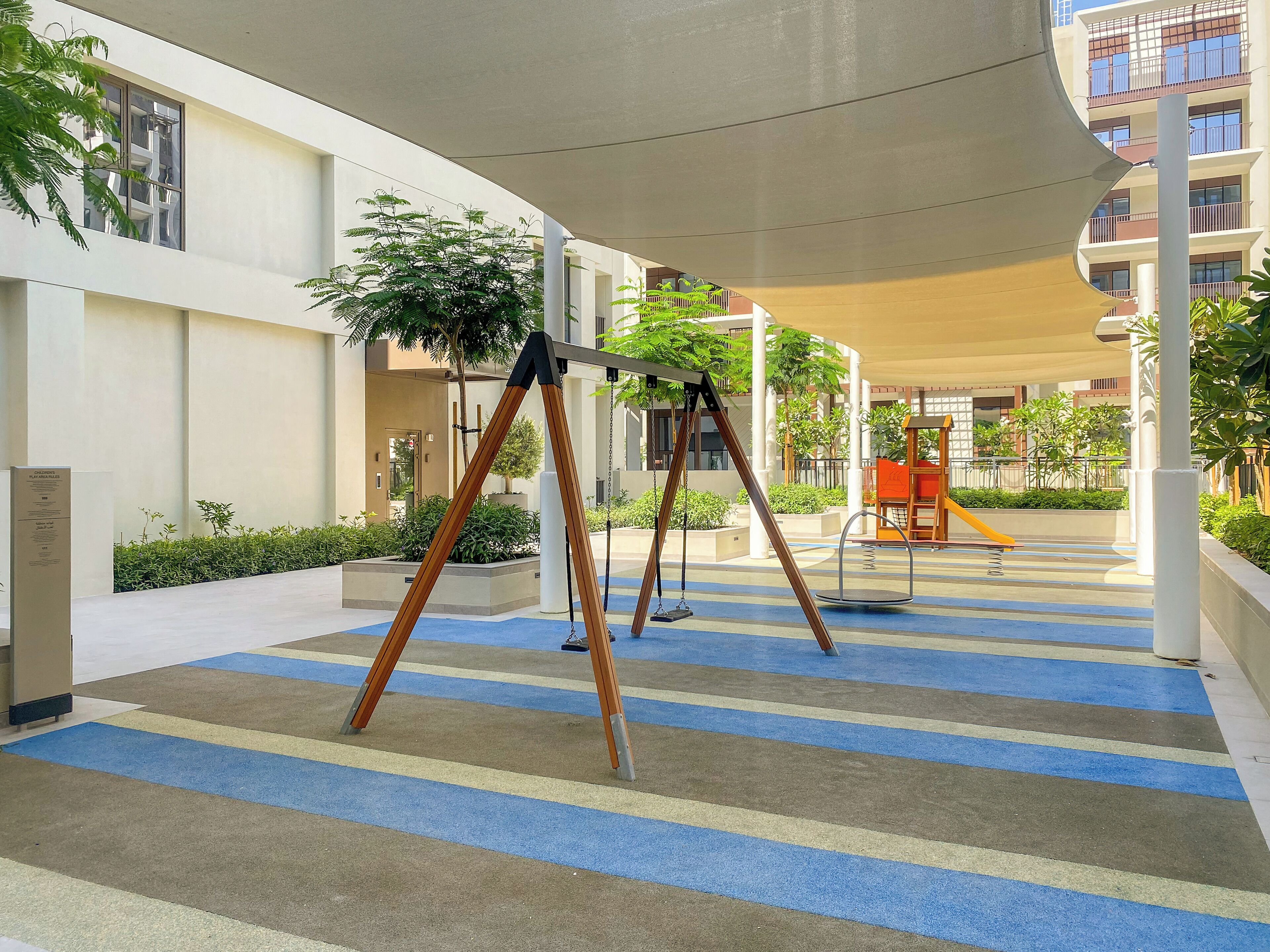 Un espacio exterior techado con un parque infantil, que incluye columpios y un tobogán, rodeado de follaje y un suelo a rayas.