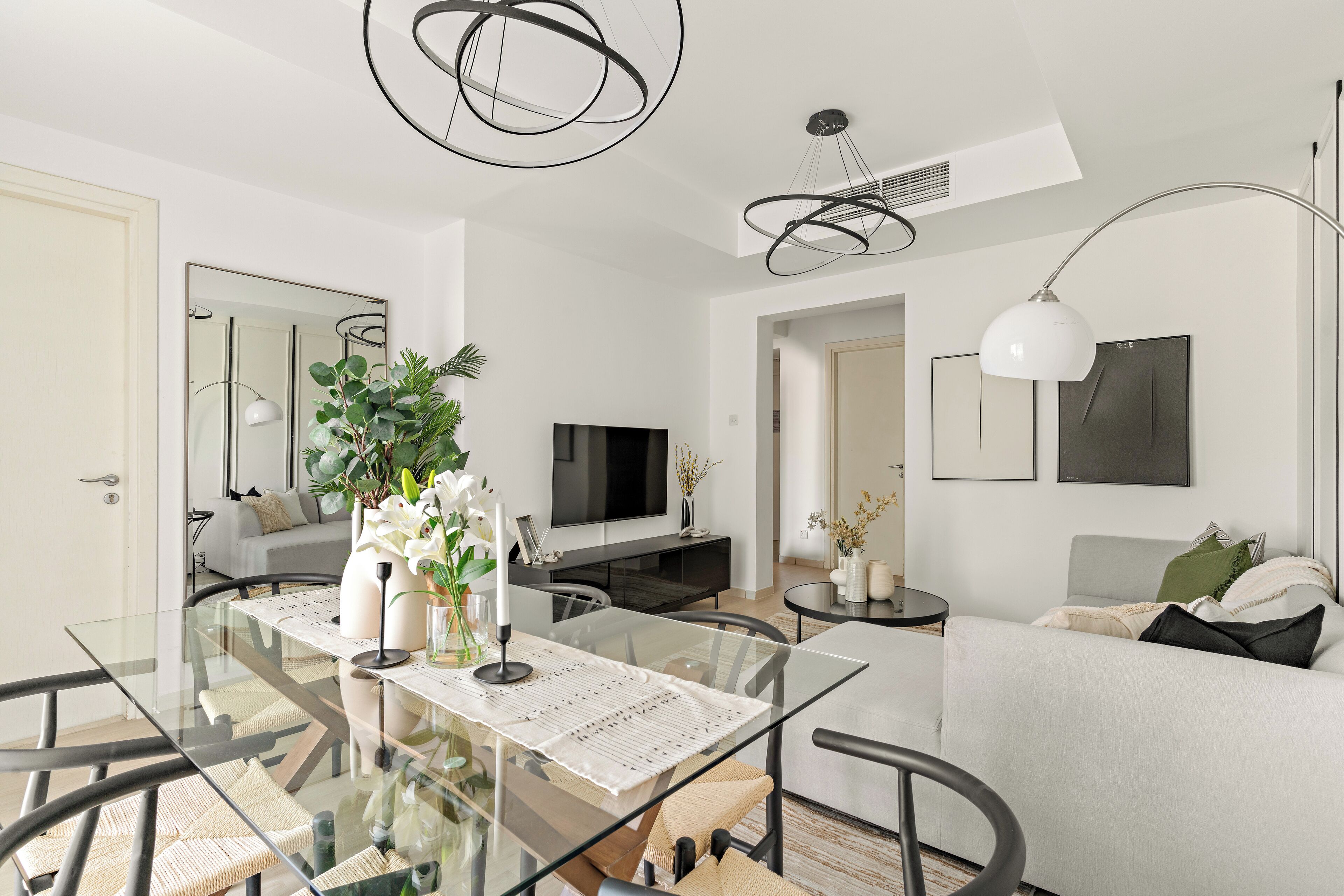 A modern living room with a glass dining table, a sofa, a TV, and a ceiling light fixture.