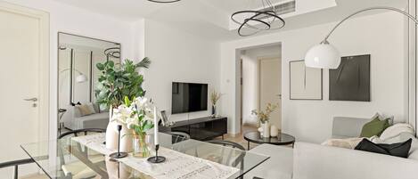 A modern living room with a glass dining table, a sofa, a TV, and a ceiling light fixture.