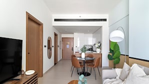 Een moderne woonkamer met een televisie, een houten tv-meubel en een bank. Er is een eethoek met een tafel en stoelen, en een keuken op de achtergrond.