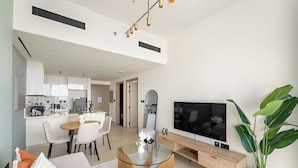 A modern living room with a glass coffee table, a sofa, a dining area with a white table and chairs, a kitchen in the background, and a large TV on a wooden stand. - Silkhaus Binghatti Corner (Dubai)