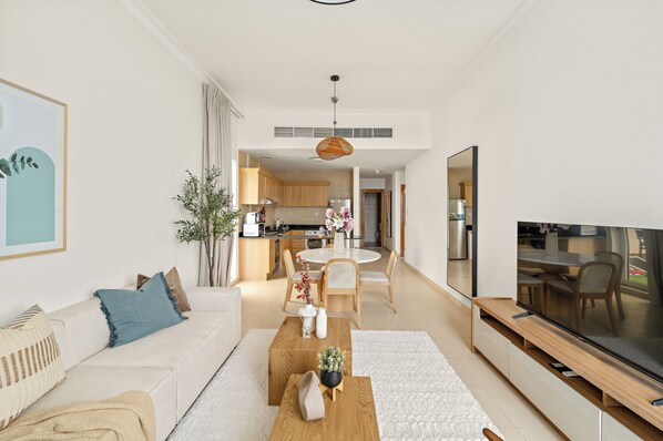 Una sala de estar moderna con sofá, mesa de centro y mueble para televisor. Se observa un área de comedor con mesa y sillas al fondo.