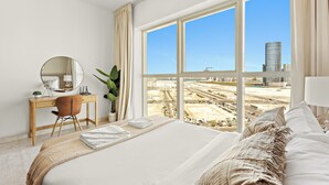 Een hotelkamer met een groot bed, een bureau met stoel, een ronde spiegel en uitzicht op een stadsgezicht door grote ramen.