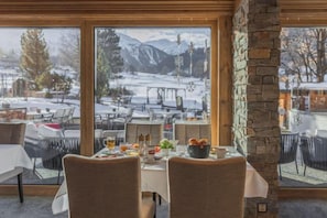 An indoor dining area with tables set for meals, large windows offering a view of a snowy outdoor scene, and a stone wall. - Sunstar Hotel Klosters (Klosters Serneus)