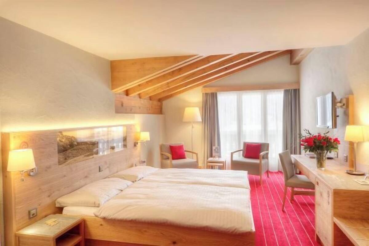 A hotel room with a wooden ceiling, a bed with white linens, two chairs, a desk, and a window with curtains.