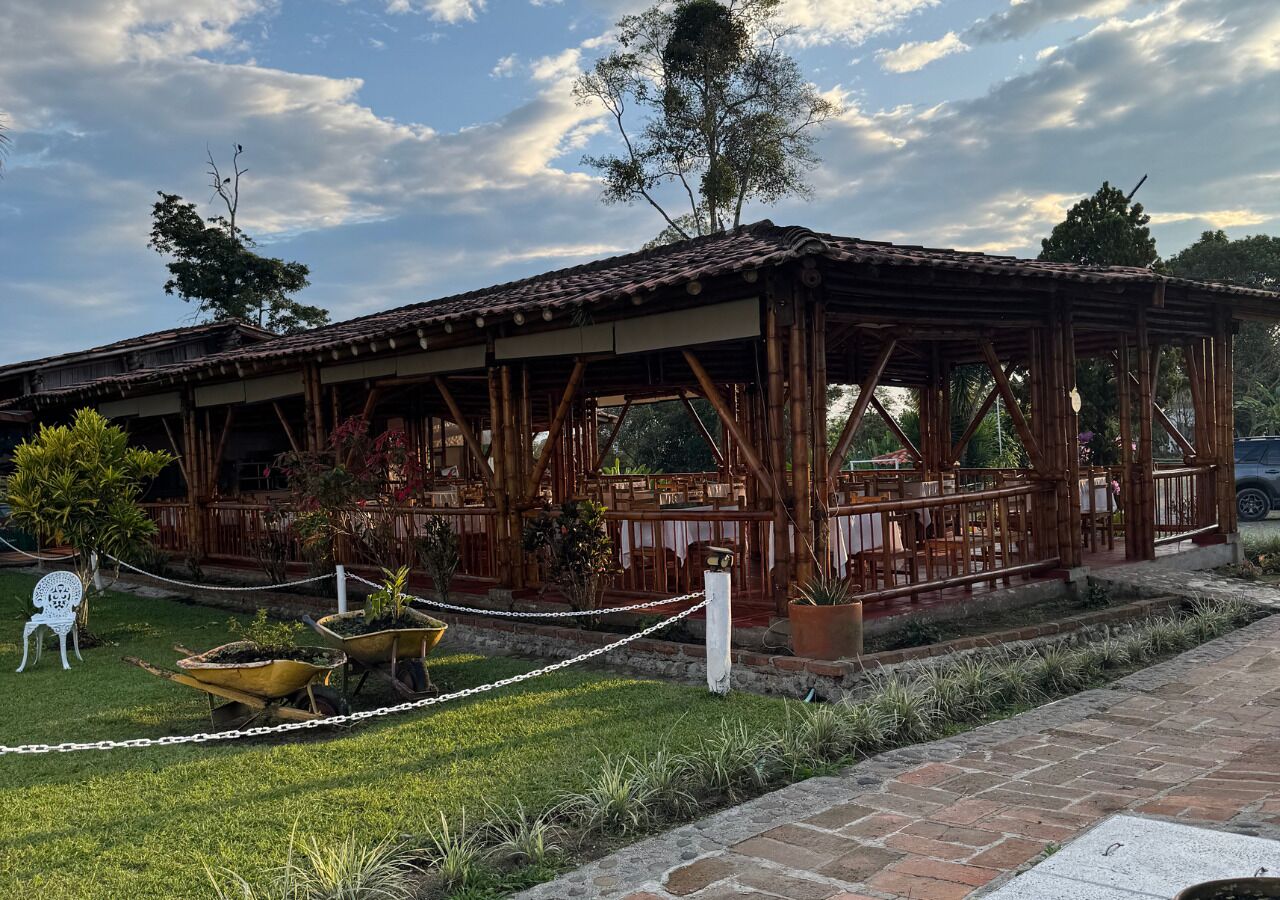 A rustic wooden pavilion with a tiled roof, surrounded by a chain-link fence, and featuring a small garden with potted plants and a wheelbarrow.