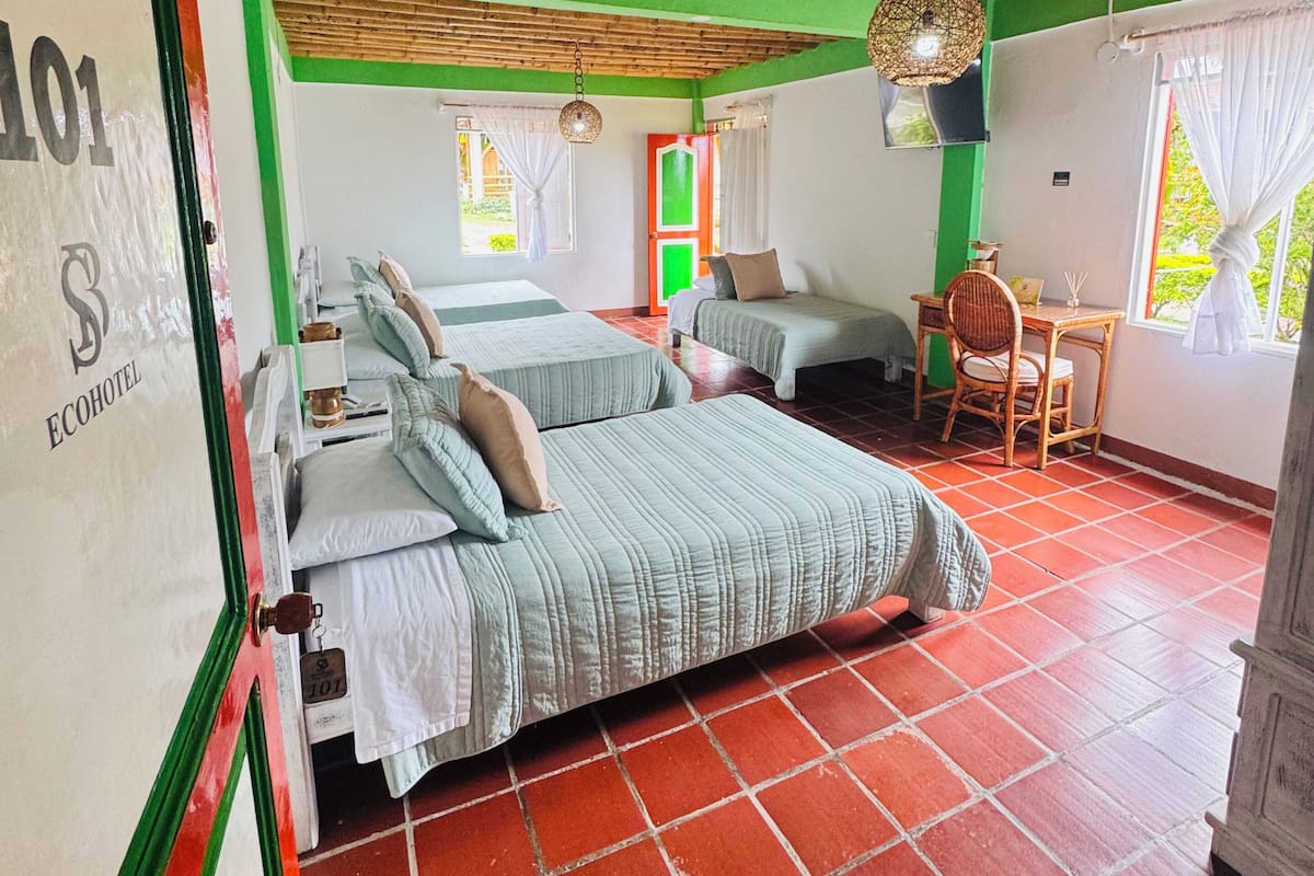 A room with a bed, a small table, and a chair. There is a window with curtains and a hanging light fixture.
