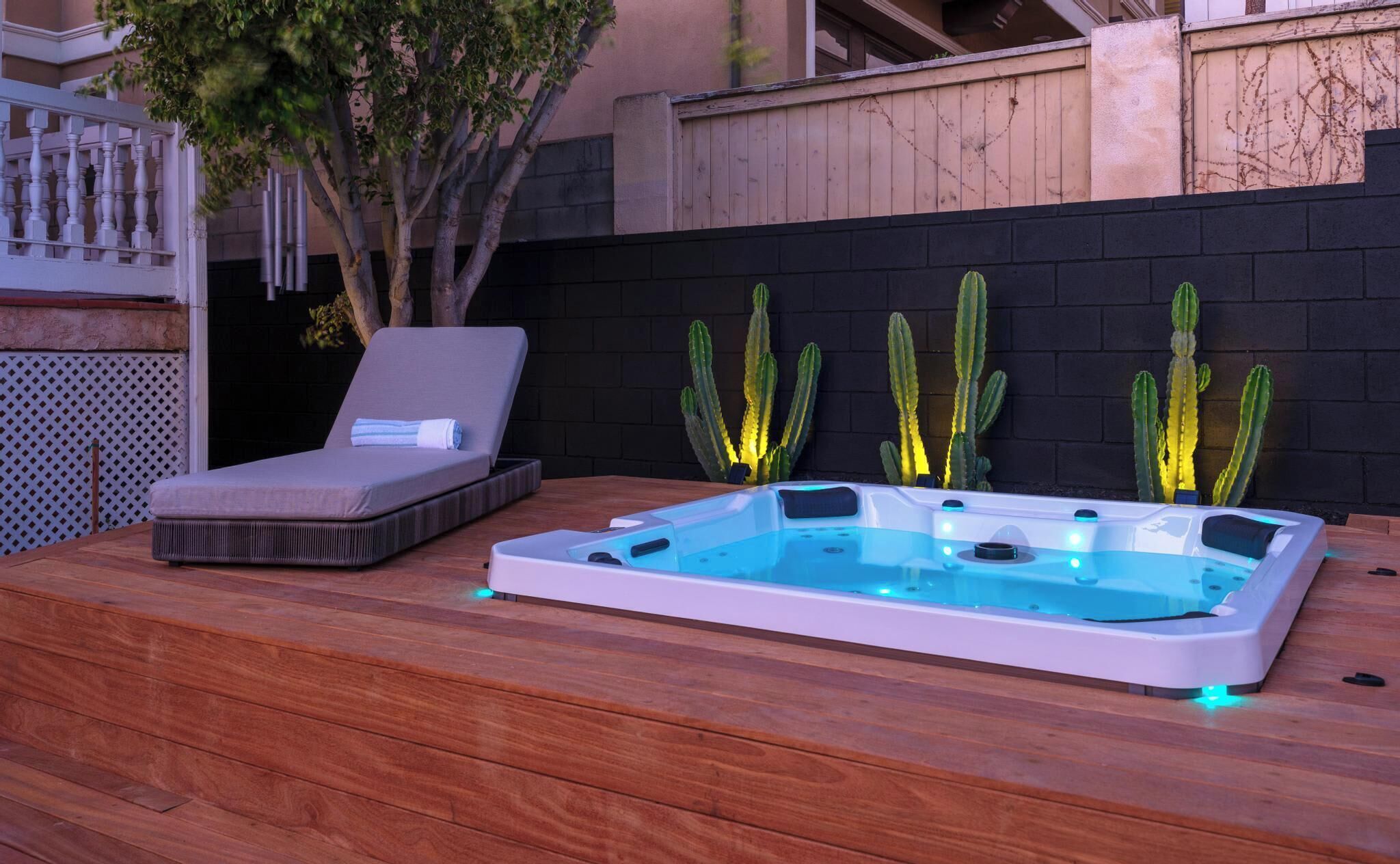 Un bain à remous avec une terrasse en bois, une chaise longue et des cactus devant un mur de briques.