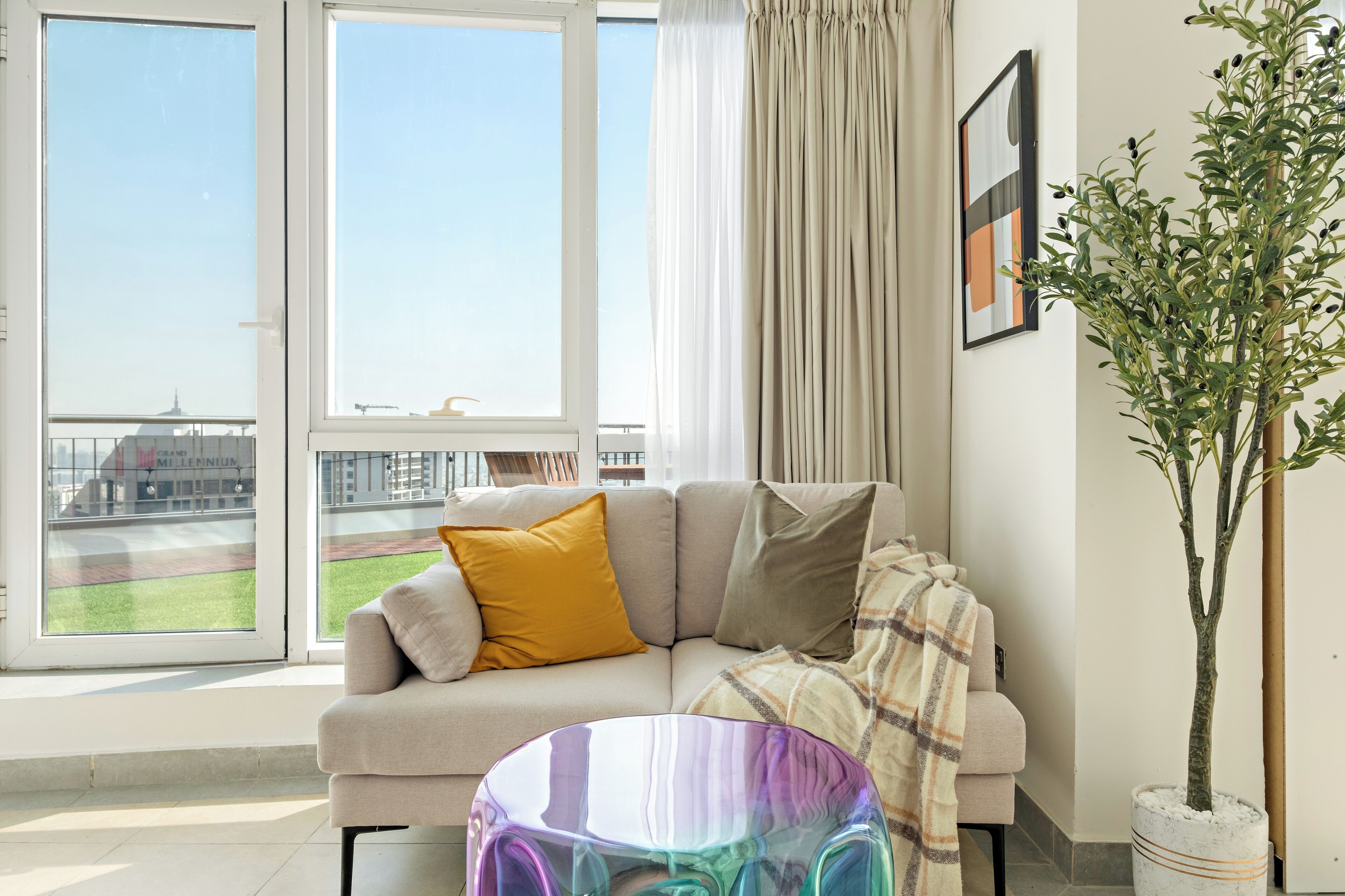 A modern living room with a sofa, a glass coffee table, a potted plant, and a view of a sports field through large windows.