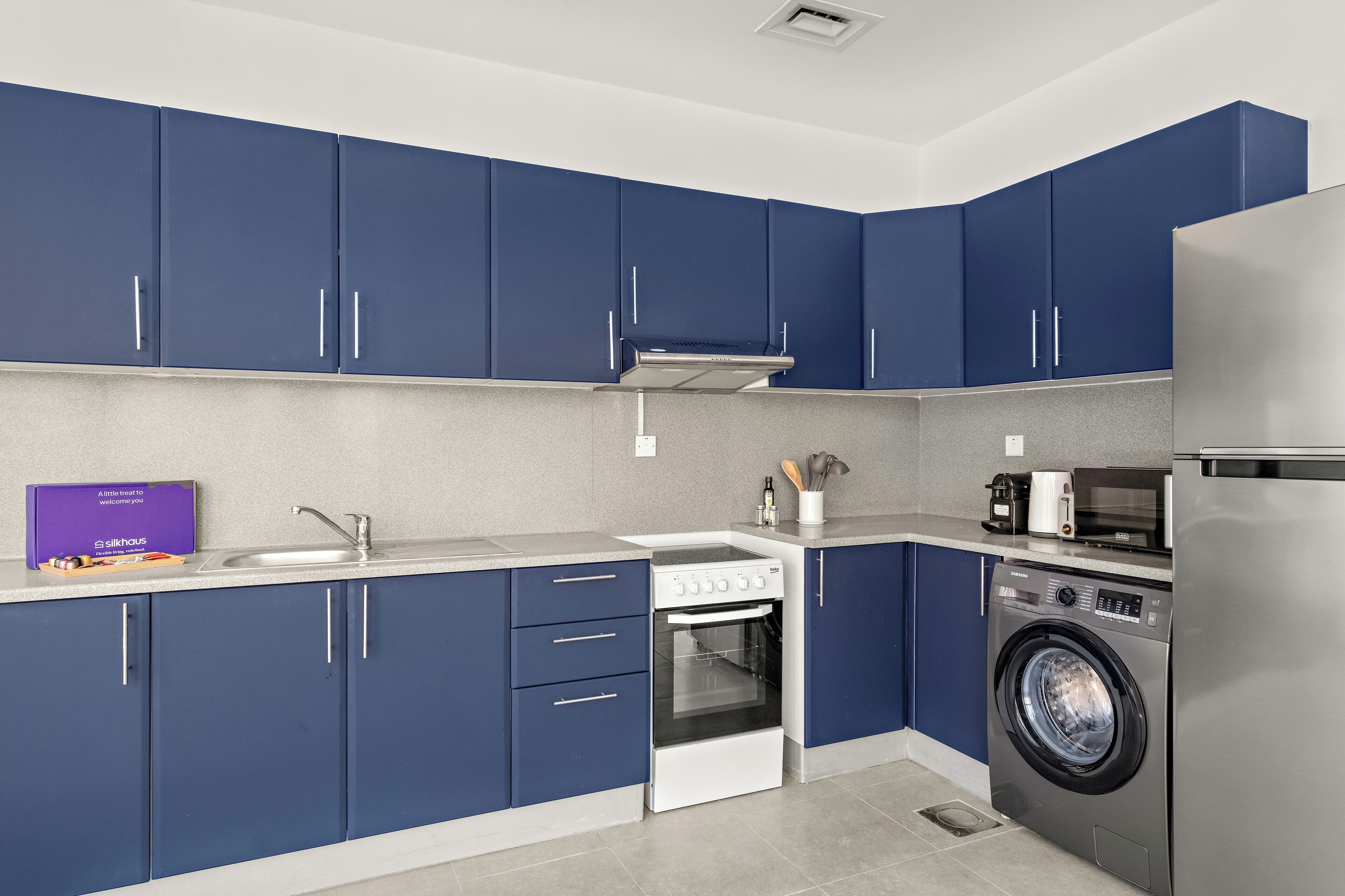 A modern kitchen with blue cabinets, a stainless steel refrigerator, a microwave, a stove, and a washing machine.