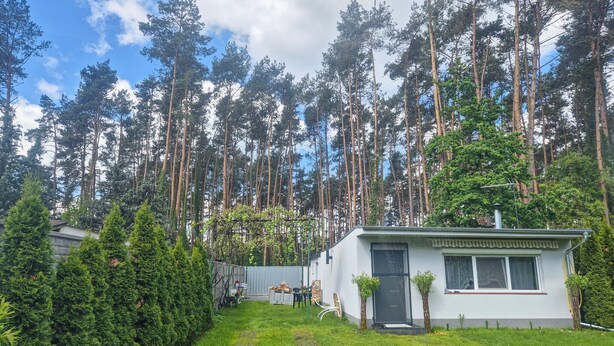 Pieni, yhden kerroksen rakennus, jonka ulkoseinät ovat valkoiset ja ovi tumma. Sen ympärillä on hyvin hoidettu puutarha ja korkeita mäntyjä.
