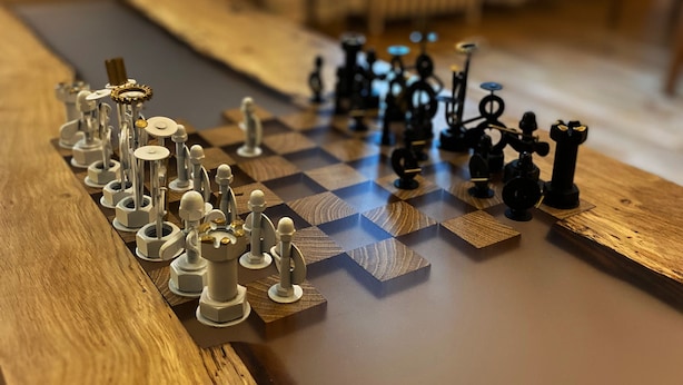 A chessboard with pieces on a wooden table, set in a room with a plant and a radiator in the background. - Le Cocon, L'Échiquier (Castres)