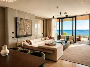 Ein modernes Wohnzimmer mit einer großen Couch, einem Couchtisch und einem Essbereich. Ein großes Fenster bietet Blick auf das Meer.