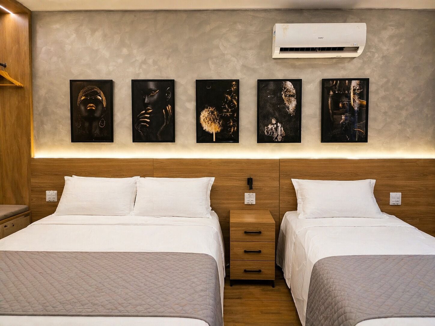 Ein Hotelzimmer mit zwei Betten, einem hölzernen Kopfteil, einer Wand-Klimaanlage und fünf gerahmten Kunstwerken.