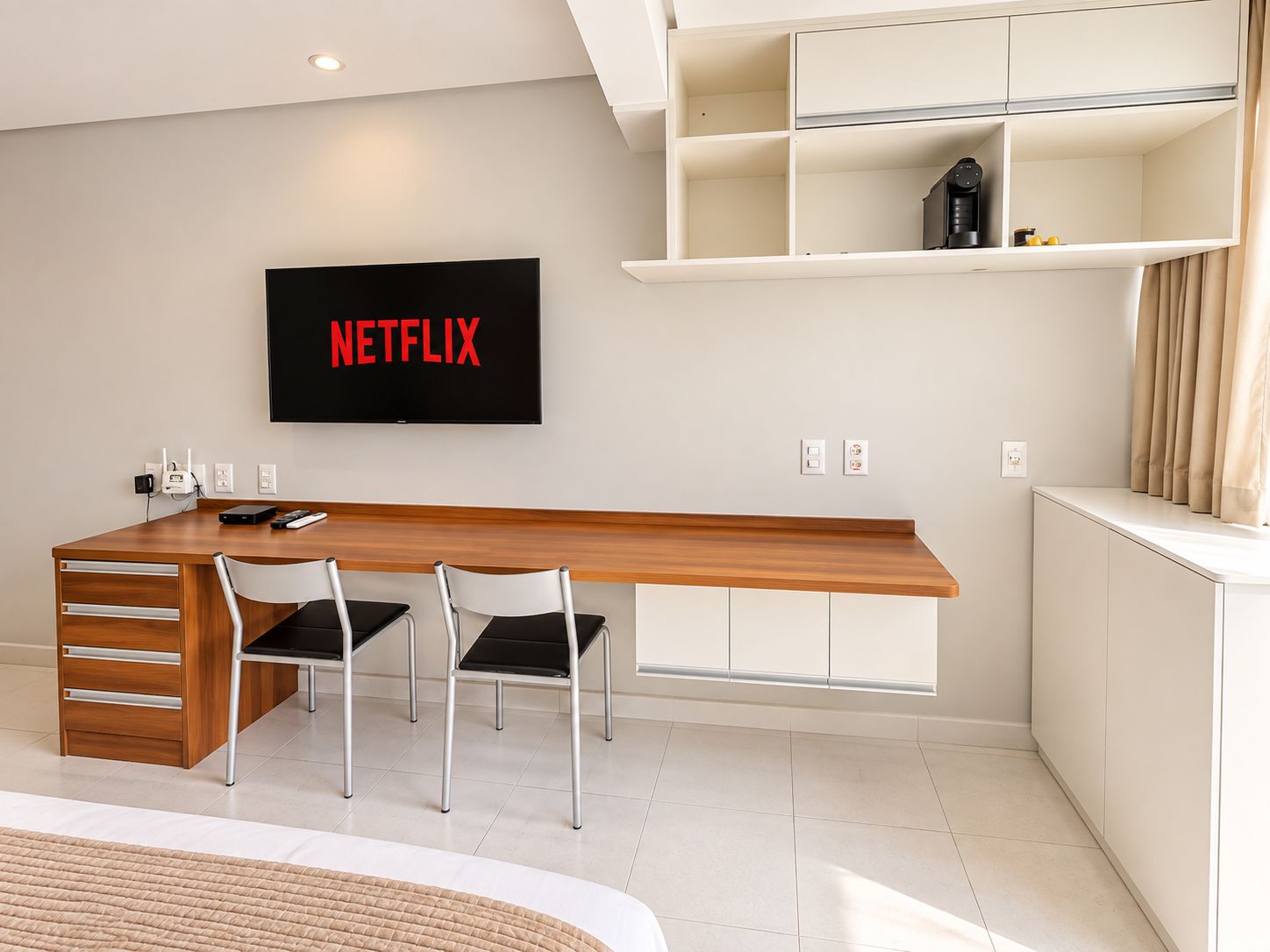 A modern hotel room with a wooden desk, two chairs, a flat-screen TV mounted on the wall, and a built-in shelf above the desk.