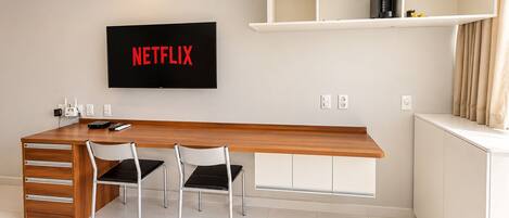 A modern hotel room with a wooden desk, two chairs, a flat-screen TV mounted on the wall, and a built-in shelf above the desk.