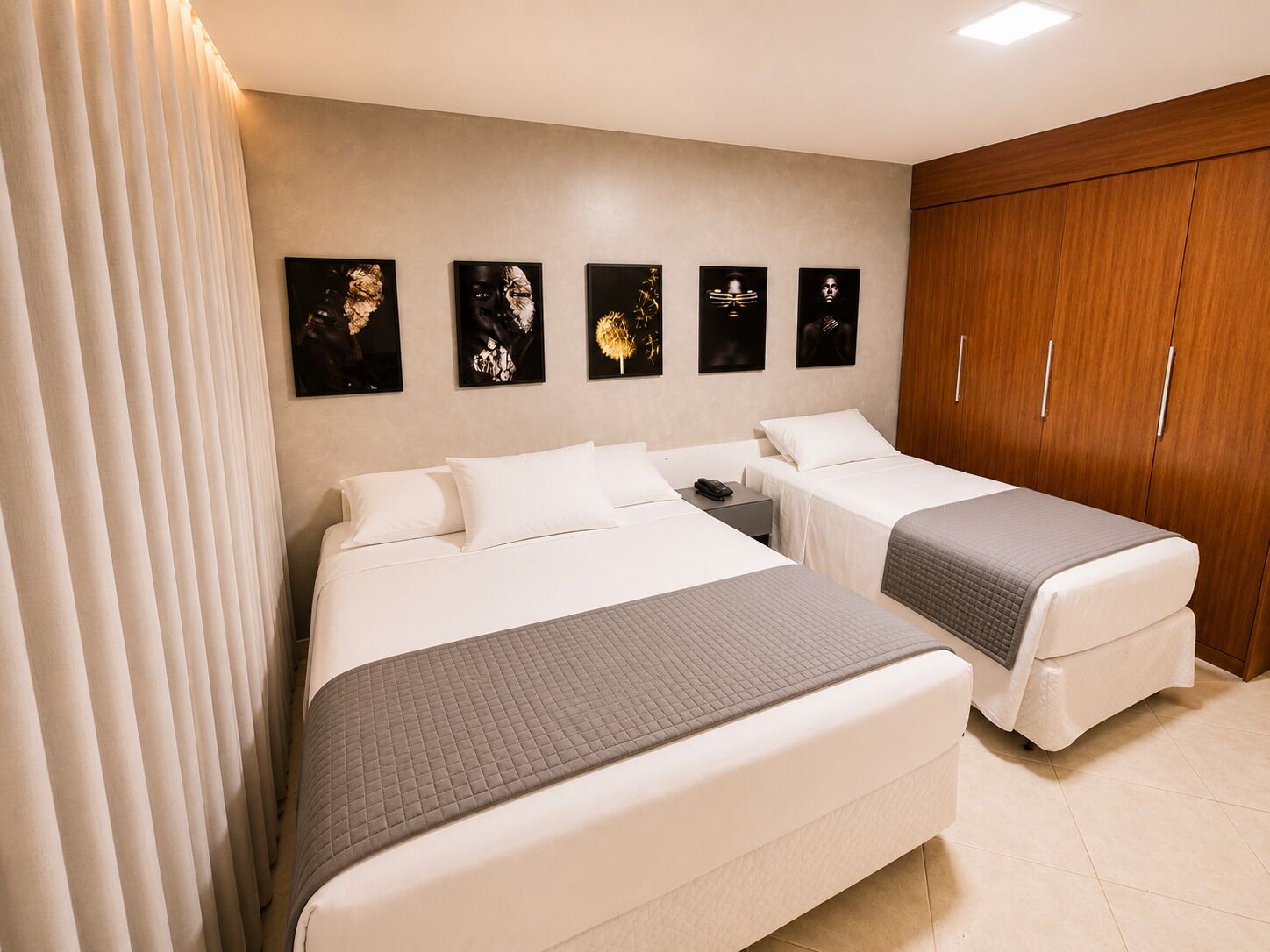 A hotel room with two beds, a wooden wardrobe, and five framed artworks on the wall.
