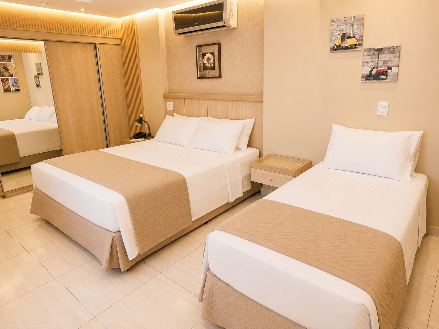 A hotel room with two beds, a wardrobe, a bedside table with a lamp, and artwork on the wall.