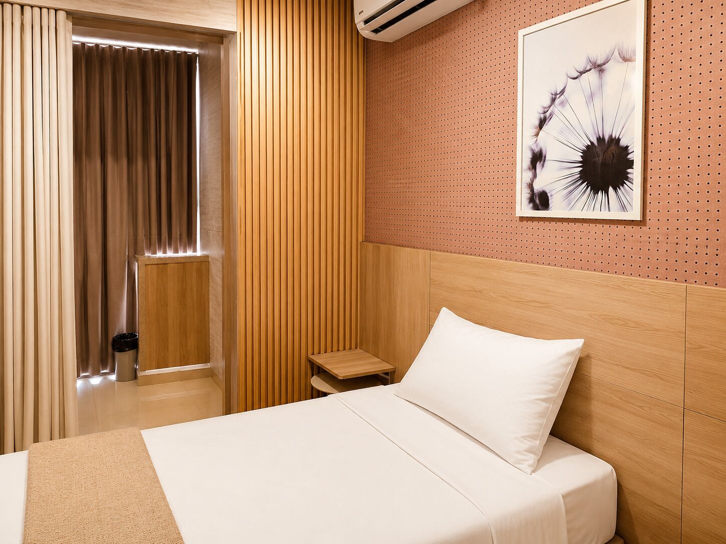 A hotel room with a bed, a nightstand, a wall-mounted air conditioner, a window with curtains, and a wall art piece.