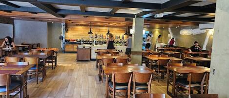 Un restaurant avec des tables et des chaises en bois, un comptoir central où travaille le chef, et un mur en briques.