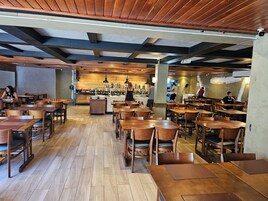 Un restaurante con mesas y sillas de madera, una barra central con un cocinero y una pared de ladrillo.