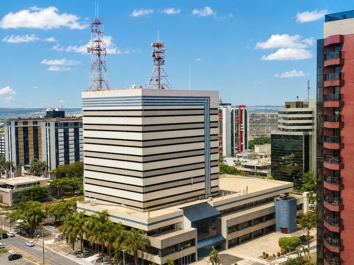 A modern building with a flat roof, surrounded by other high-rise buildings, featuring a clear sky and some palm trees.