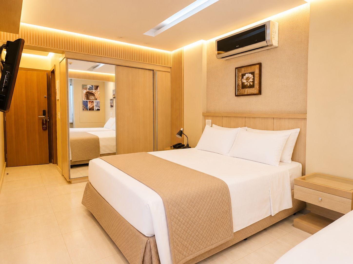 A hotel room with a large bed, a bedside table, an air conditioning unit, a wardrobe, and a television.