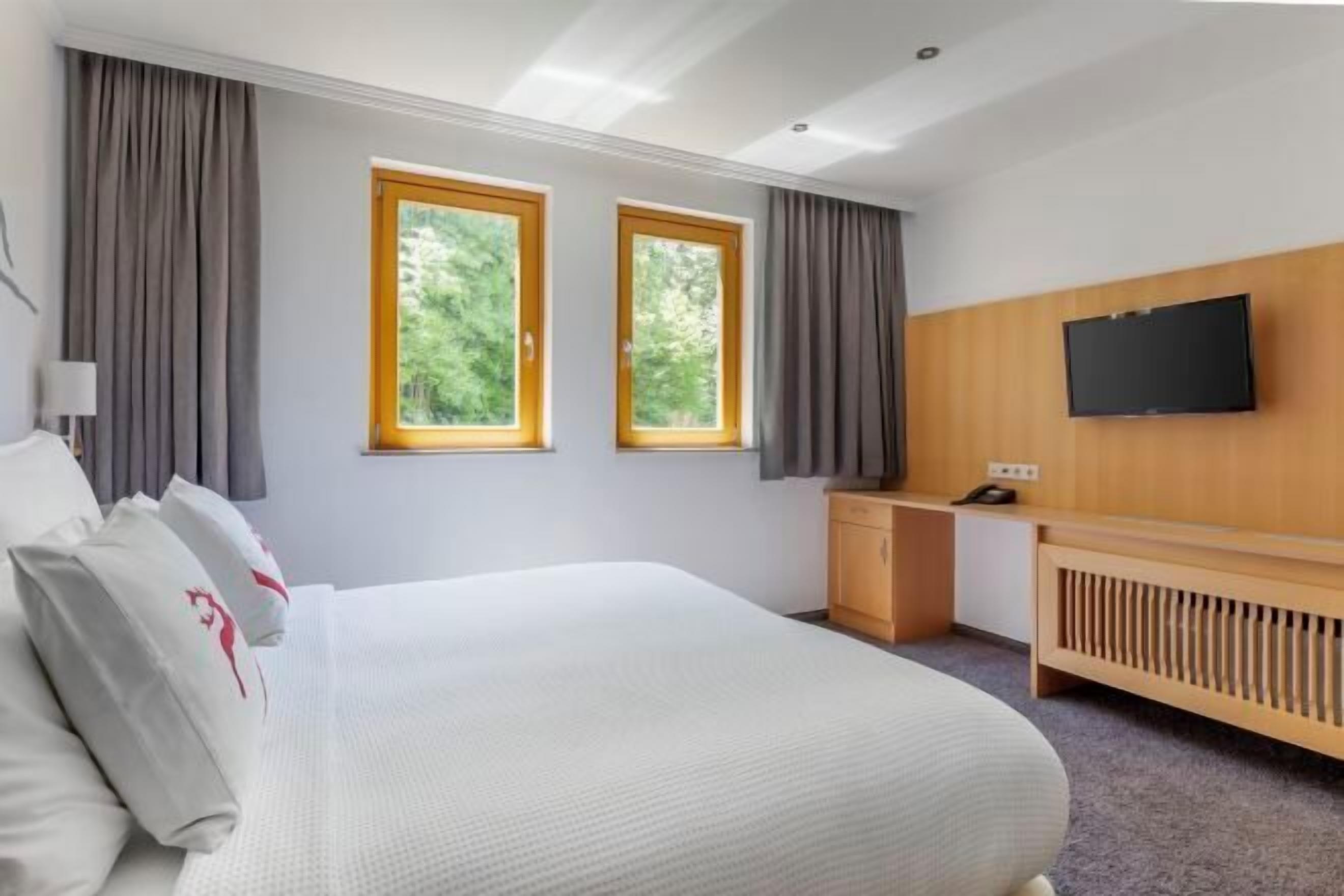 Une chambre d’hôtel moderne équipée d’un grand lit, d’un bureau, d’une télévision et de deux fenêtres avec des rideaux.