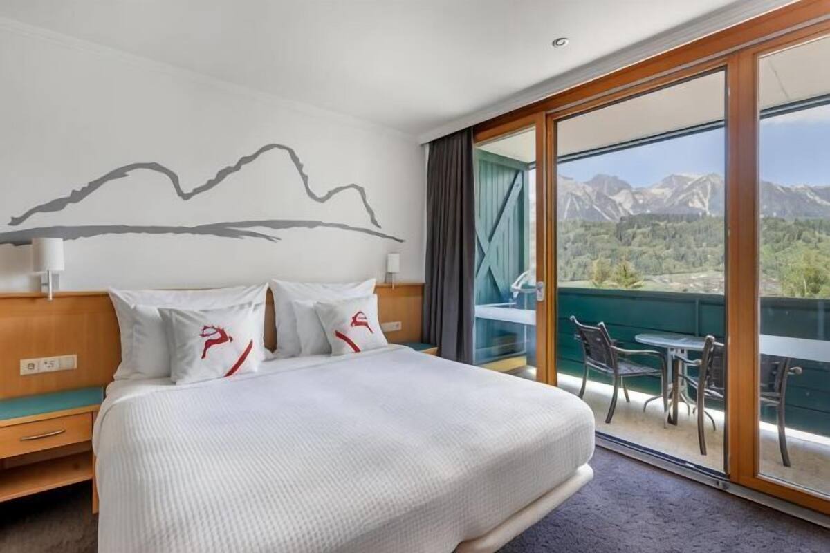 A hotel room with a bed, a mountain view, and a balcony with chairs.