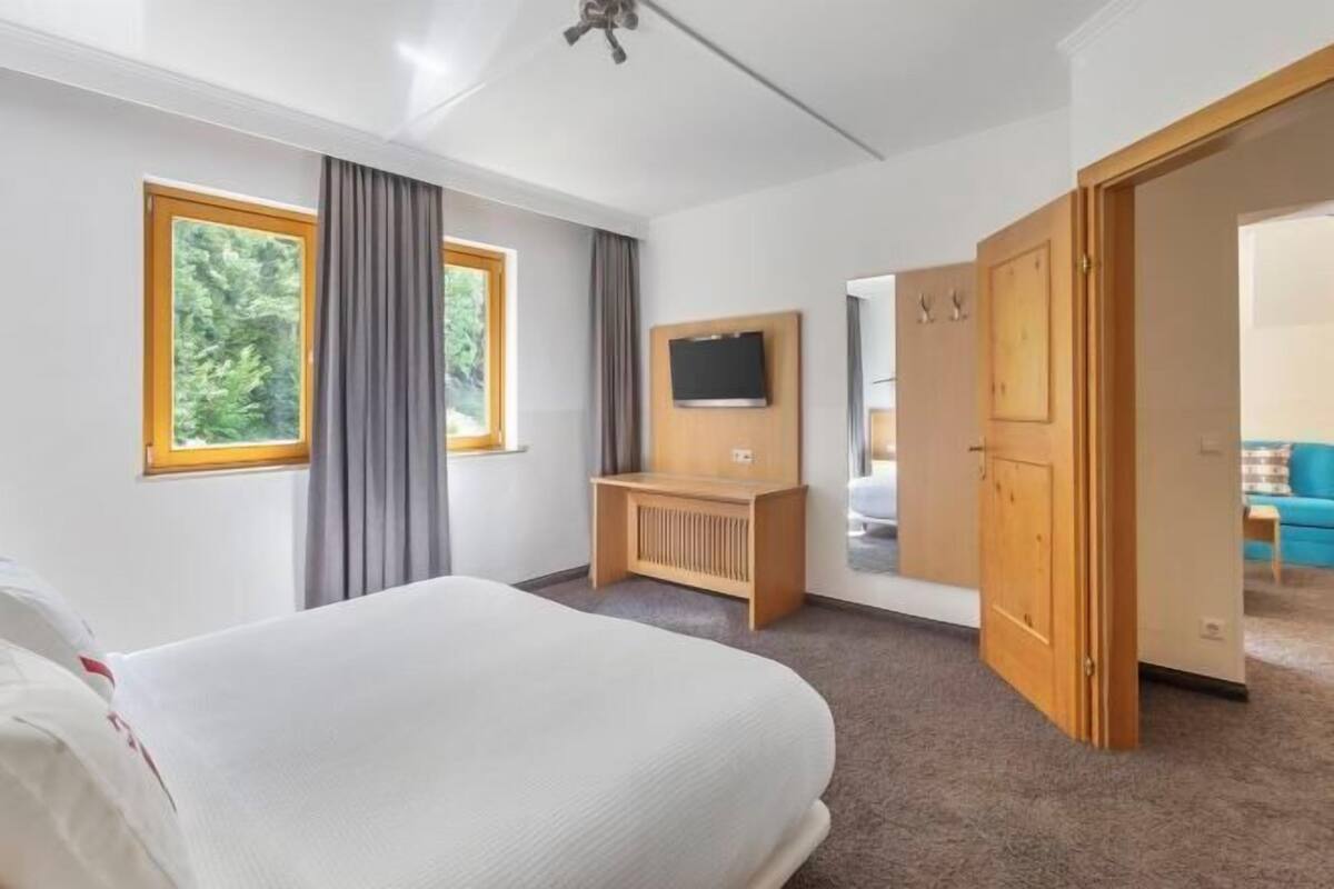 A hotel room with a bed, a TV mounted on the wall, a wooden desk, and a door leading to another room.