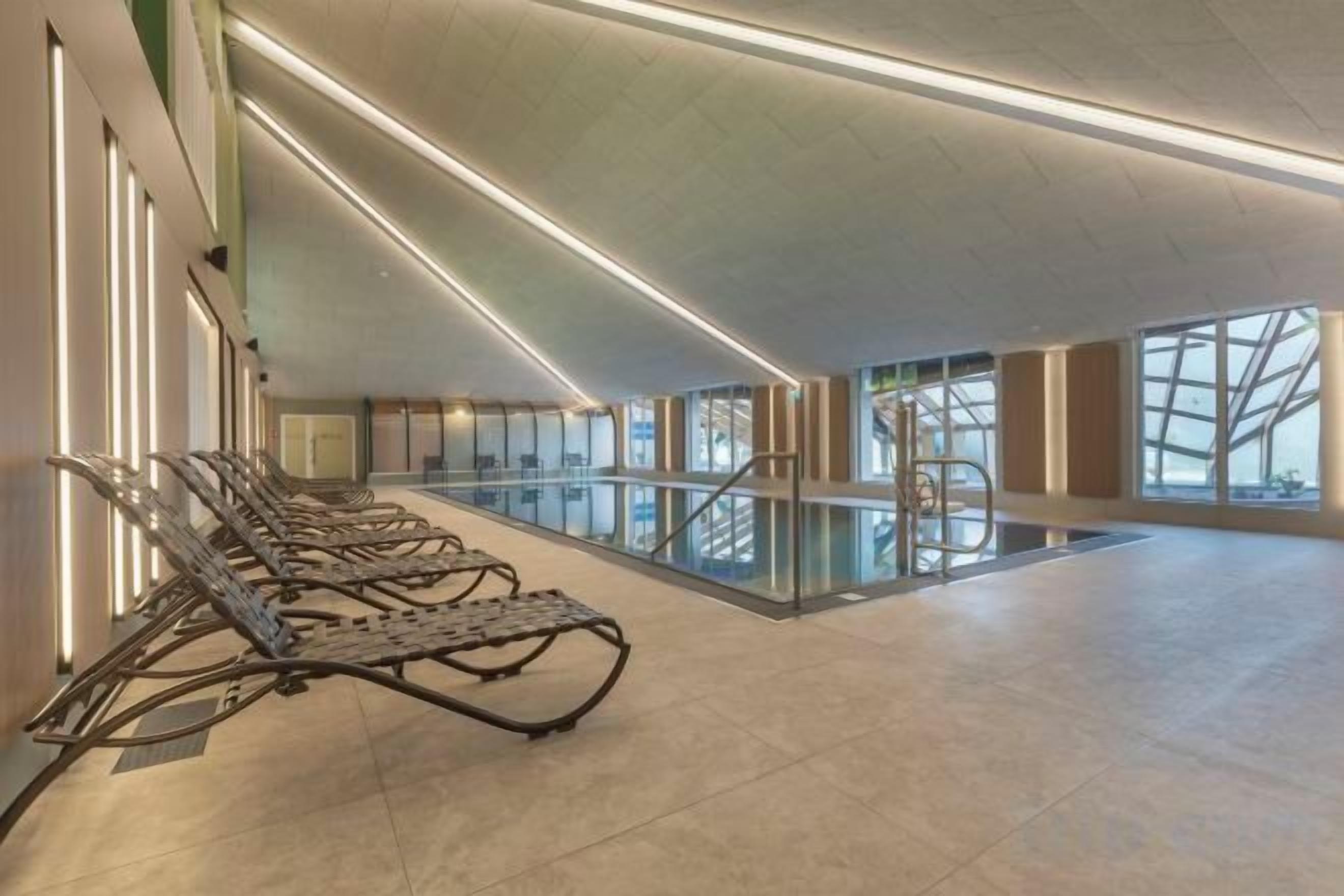 Un área de piscina interior moderna con techos iluminados por el sol, una vista despejada del cielo a través de amplias ventanas y una fila de sillones de sol.