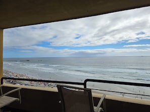 Vista de una playa desde un balcón con sillas, un muelle y un barco a lo lejos.
