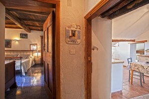 Une pièce chaleureuse avec une porte en bois, un lit recouvert d’une courtepointe blanche, une petite table avec une nappe à motifs et un coin cuisine visible à travers une porte ouverte.