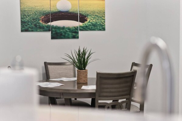 Et spiseområde med et rundt bord, to stole, en potteplante og et indrammet kunstværk med golfmotiver på væggen.