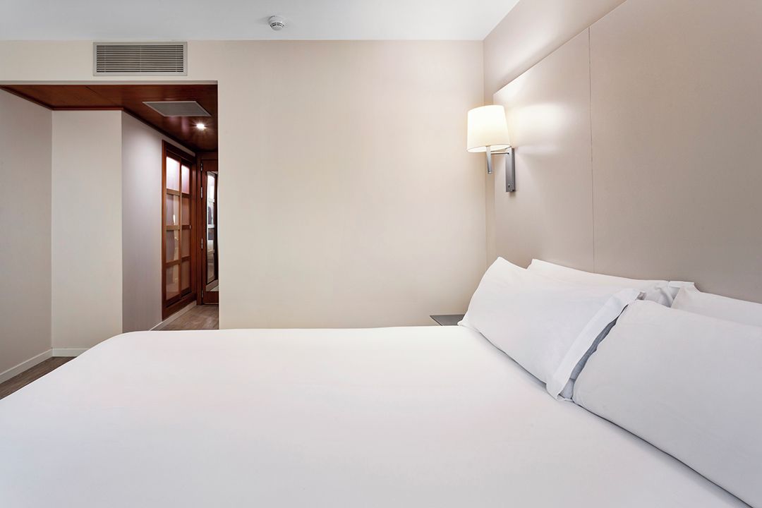 Een hotelkamer met een groot bed, een wandlamp en een houten deur naar een gang.