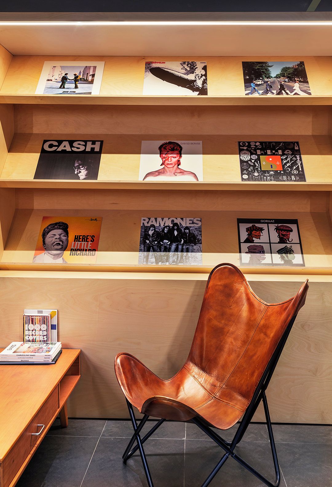 A modern interior with a wooden shelf displaying various album covers, a leather chair, and a small table with magazines.
