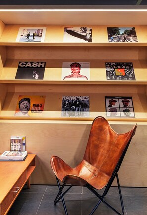 A modern interior with a wooden shelf displaying various album covers, a leather chair, and a small table with magazines. - B&B Hotel Castellón (Castelló de la Plana)